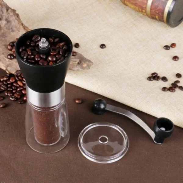 Black hand crank coffee grinder filled with beans, with the removable steel handle and clear lid beside it